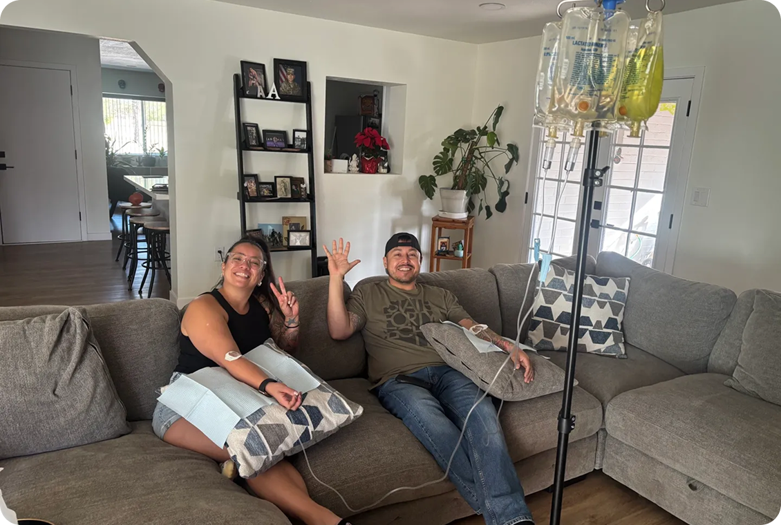 Two people sitting on a couch waving at the camera in a cozy living room.