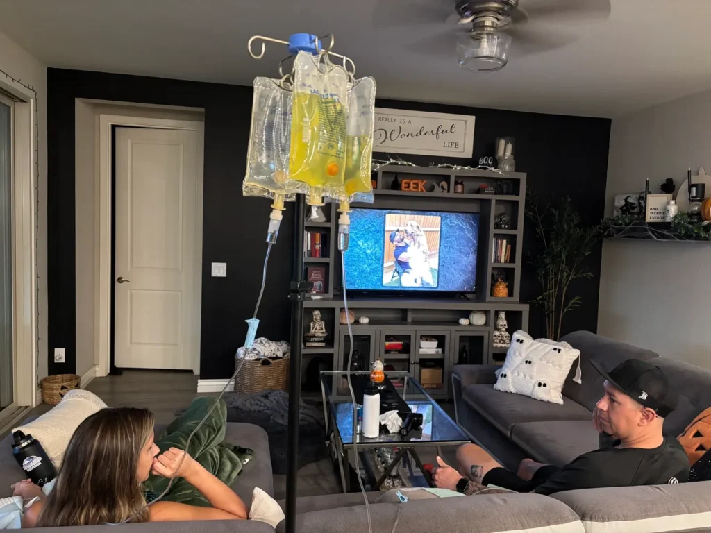Two people relaxing in a living room with a TV and an IV drip stand.
